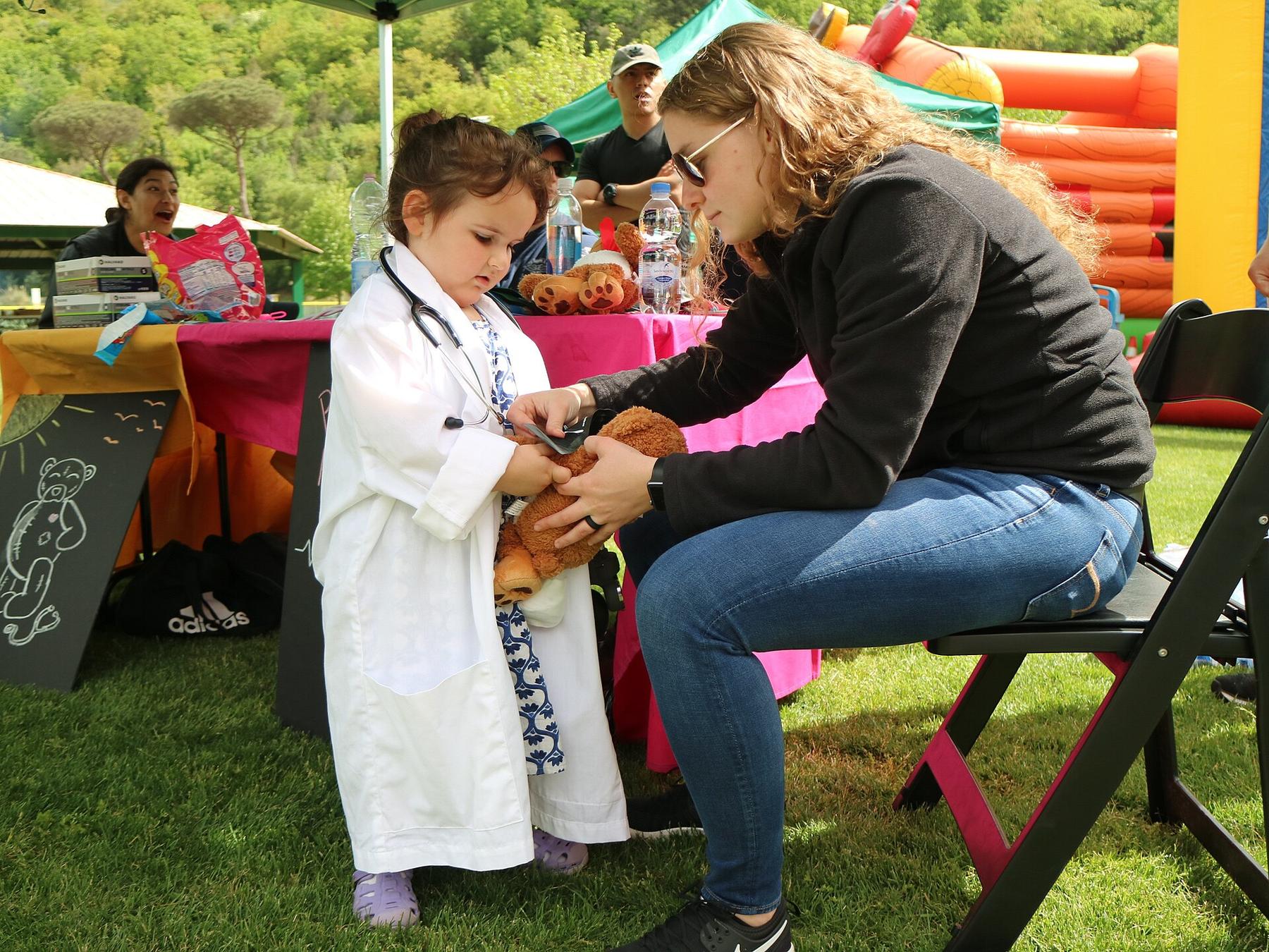 A child in a doctor's coat checking a teddy bear at an outdoor fair